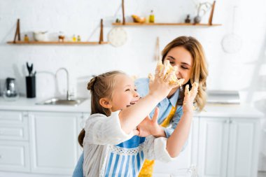 Gülümseyen anne ve kız ellerinde hamurla mutfakta oynuyorlar. 