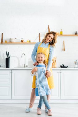 Gülümseyen anne kızını kucaklıyor ve mutfakta kameraya bakıyor. 
