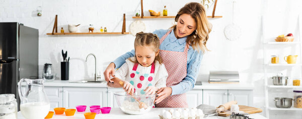 Beautiful attractive mother standing behind cute daughter while kneading dough, panoramic shot