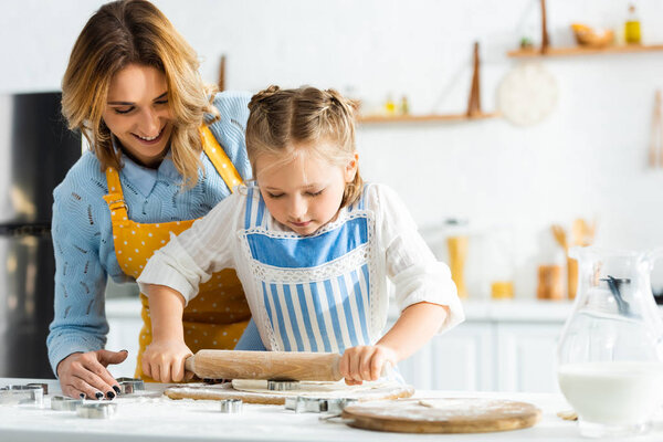 smiling mother and daughter cooking with rolling pin in kitchen 