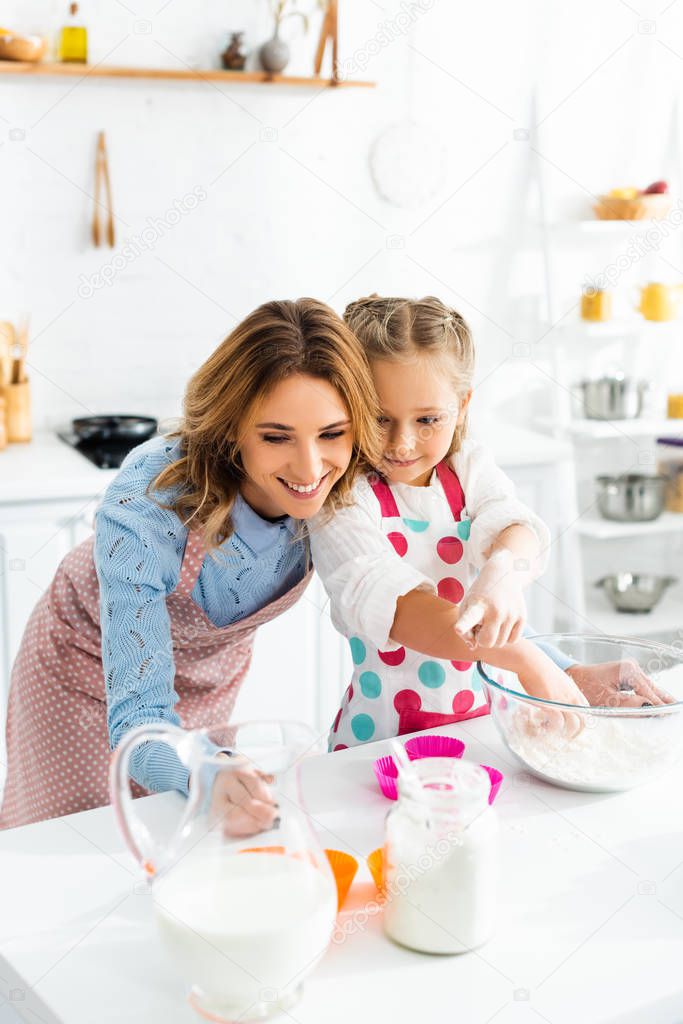 Attractive mother and cute daughter preparing cupcakes with ingredients including milk, flour in kitchen