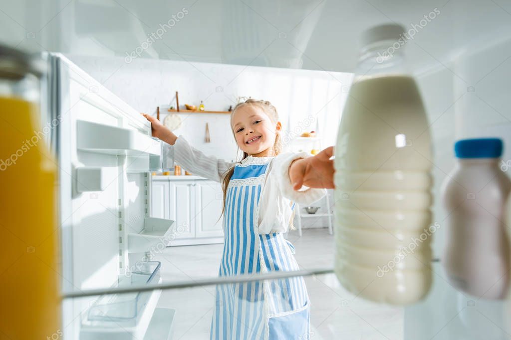 selective focus of smiling kid taking bottle with milk from fridge 
