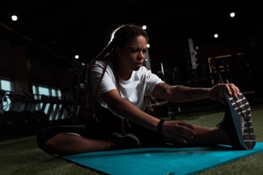 Afrikalı Amerikalı kadın spor salonunda oturmuş spor paspasının üzerinde geriniyor.