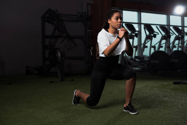 Young adult african american woman clenching hands and doing lunges in gym