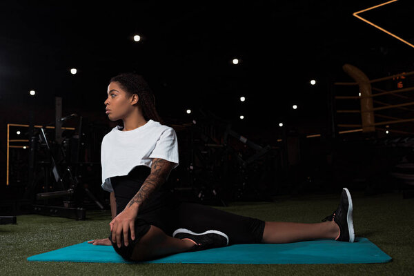 Attractive african american woman sitting and stretching on fitness mat in gym