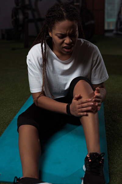 African american woman suffering from pain in leg, sitting on fitness mat