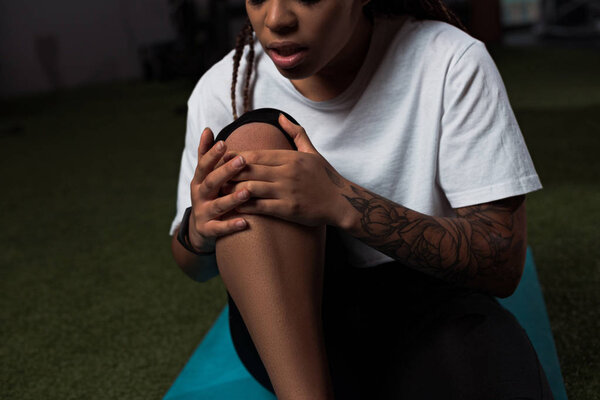 Cropped view of african american woman suffering from pain in knee, sitting on fitness mat