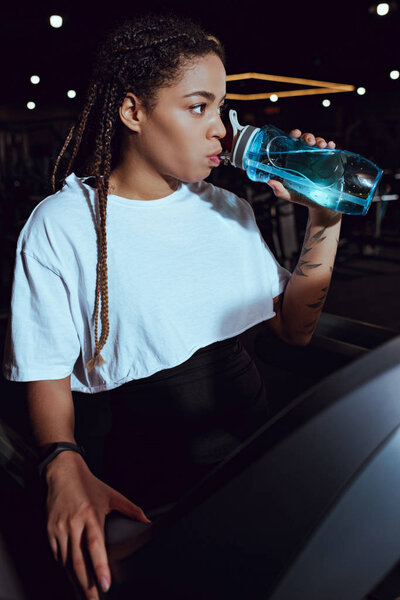 Selective focus of african american woman drinking water from sports bottle on treadmill