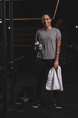 happy african american girl with tattoo holding sports bottle with water and standing in gym 