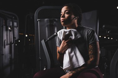 tattooed african american woman listening music and holding towel in gym 