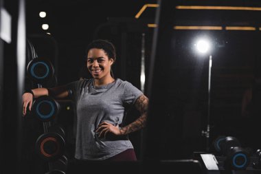 selective focus of happy african american girl with dreadlocks standing with hand on hip near barbells in gym 
