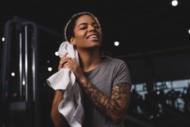 tattooed african american girl wiping sweat with towel and smiling n gym 