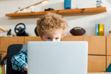 Laptop kullanan zeki ve kıvırcık çocuğun seçici odağı 