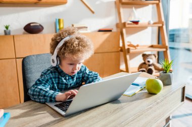 Kıvırcık çocuk, kablosuz kulaklıkla müzik dinlerken elmanın yanında dizüstü bilgisayar kullanıyor. 