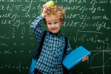 Matematiksel formüllerle dolu karatahtanın yanında elma ve kitap tutan mutlu çocuk. 