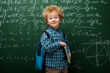 Matematiksel formüllerle karatahtanın yanında mutlu çocuk kitabı tutuyor. 