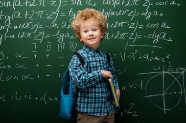 happy child holding book near chalkboard with mathematical formulas  clipart