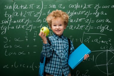 Matematiksel formüllerle dolu karatahtanın yanında elma ve kitap tutan neşeli çocuk. 