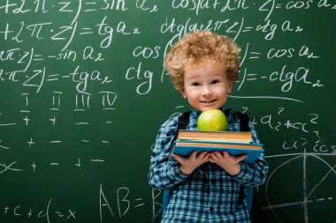 Matematiksel formüllerle dolu karatahtanın yanında kitap ve elma tutan neşeli çocuk. 