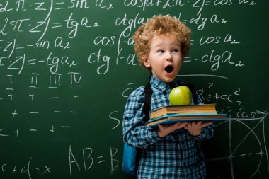 Şaşırmış çocuk elinde matematik formüllü karatahtanın yanında kitap ve elma tutuyor. 