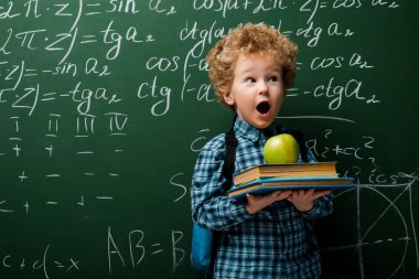 surprised kid holding books and apple near chalkboard with mathematical formulas  clipart