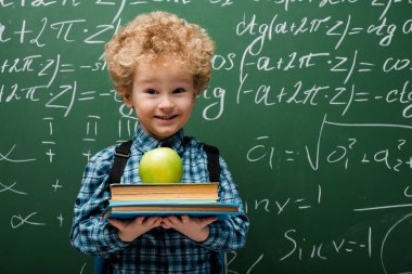 curly kid holding books and apple near chalkboard with mathematical formulas  clipart
