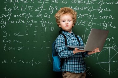 Matematiksel formüllerle karatahtanın yanında dizüstü bilgisayar tutan kıvırcık çocuk. 