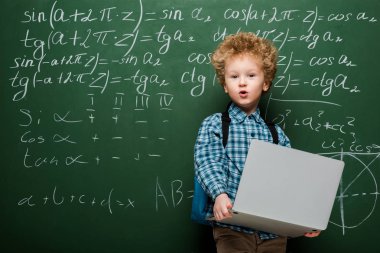 Şaşırmış çocuk elinde matematiksel formüllerle karatahtanın yanında dizüstü bilgisayar tutuyor. 