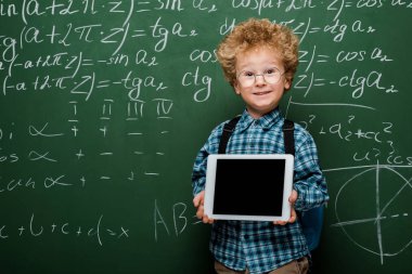 Gözlüklü mutlu ve zeki çocuk elinde boş ekranlı dijital tablet, matematiksel formüllü karatahtanın yanında. 