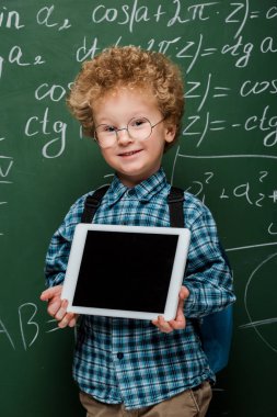 Gözlüklü neşeli çocuk elinde dijital tabletle kara tahtanın yanında matematik formülleri olan boş bir ekran tutuyor. 