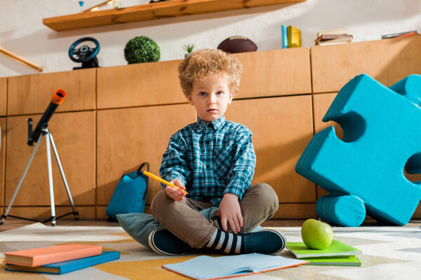 Curly and smart kid holding pen and sitting on floor near books
 