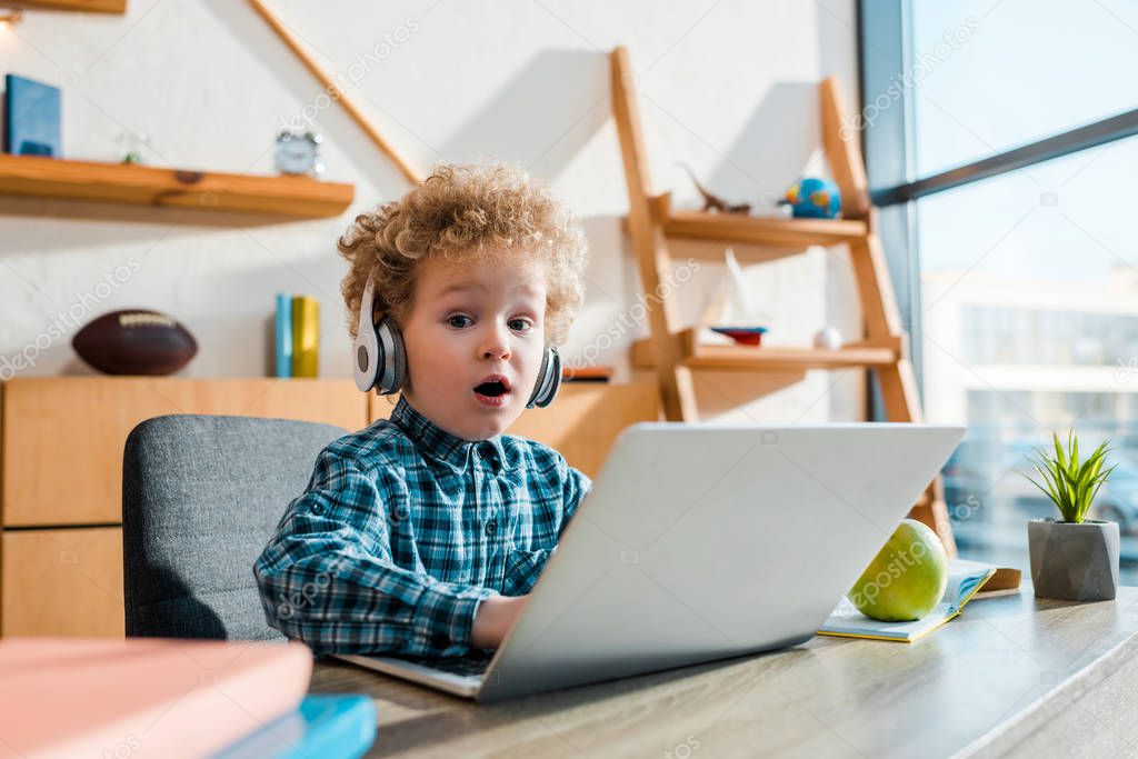 selective focus of surprised kid typing on laptop near apple while listening music in wireless headphones 