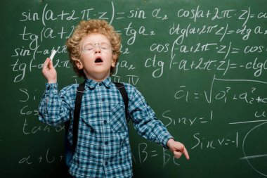 Matematiksel formüllerle kara tahtanın yanında hapşıran gözlüklü kıvırcık çocuk. 