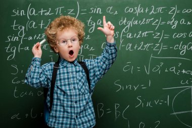 Matematiksel formüllerle kara tahtanın yanında el kol hareketi yapan gözlüklü kıvırcık çocuk. 