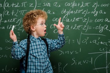 surprised kid in glasses gesturing while holding chalk near chalkboard with mathematical formulas  clipart