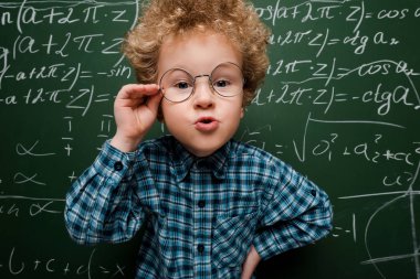smart kid touching glasses and standing with hand on hip near chalkboard  clipart
