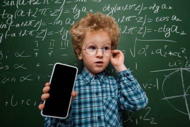Gözlüklü akıllı çocuk elinde beyaz ekranlı akıllı telefon tutuyor. Matematiksel formüllü karatahtanın yanında. 