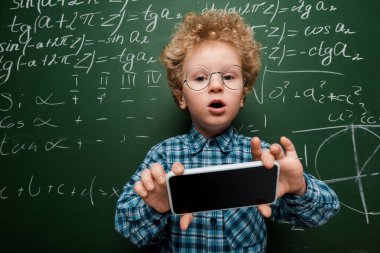 Gözlüklü akıllı çocuk elinde beyaz ekranlı akıllı telefon tutuyor. Matematiksel formüllü karatahtanın yanında. 