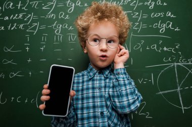 Akıllı çocuk elinde boş ekranlı akıllı telefon ve karatahtanın yanında matematik formüllü gözlüklere dokunuyor. 