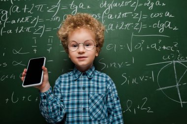 Akıllı çocuk elinde beyaz ekranlı akıllı telefonu matematik formüllü karatahtanın yanında tutuyor. 