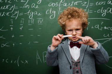 smart child in suit with bow tie pointing with finger at chalk near chalkboard with mathematical formulas  clipart