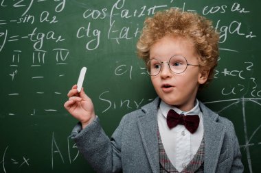smart child in suit and bow tie holding chalk near chalkboard with mathematical formulas  clipart