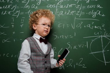 Takım elbiseli ve papyonlu hoşnutsuz çocuk elinde beyaz ekranlı, matematiksel formüllü akıllı telefon tutuyor. 
