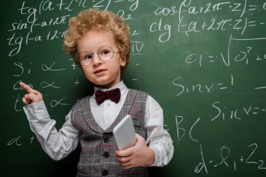 smart child in suit and bow tie holding smartphone and pointing with finger near chalkboard with mathematical formulas  clipart