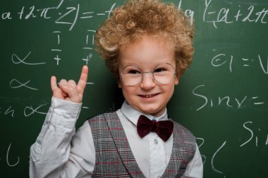 Takım elbiseli ve papyonlu mutlu ve zeki çocuk matematiksel formüllü karatahtanın yanında kaya işareti gösteriyor. 