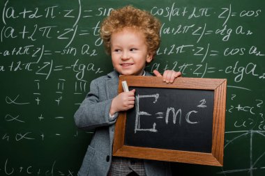 cheerful kid in suit and bow tie holding small blackboard with formula near chalkboard  clipart