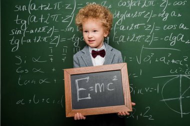 smart child in suit and bow tie holding small blackboard with formula near chalkboard  clipart