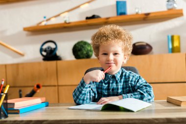 happy and curly kid holding pen near notebook and books on desk  clipart