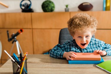 selective focus of excited kid sitting near books on table  clipart