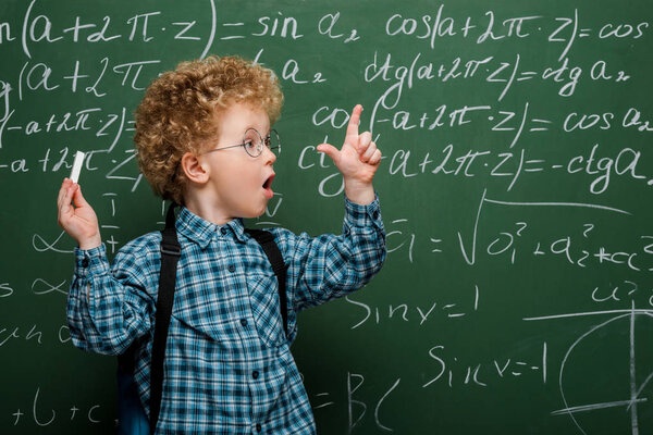 surprised kid in glasses gesturing while holding chalk near chalkboard with mathematical formulas 